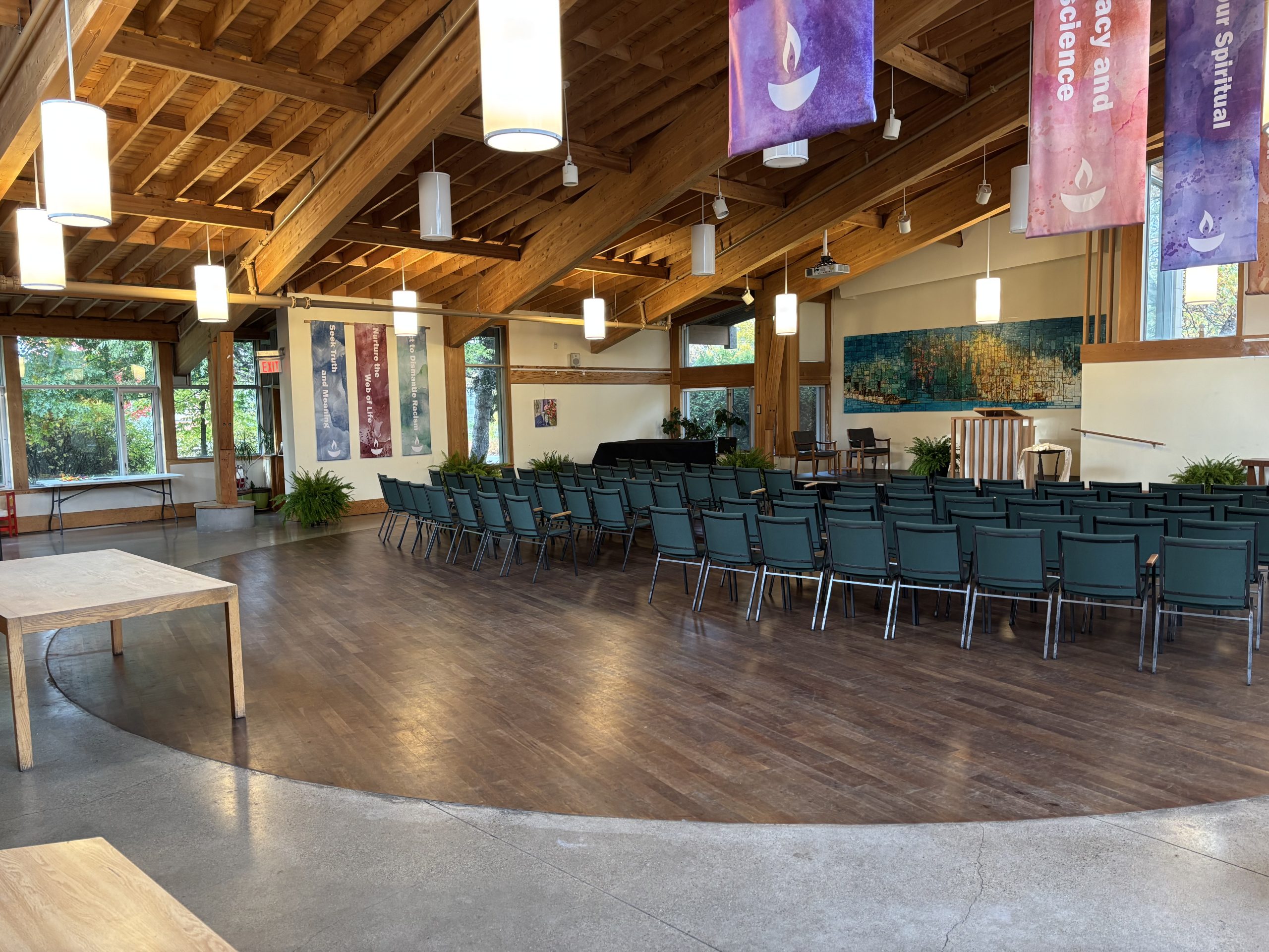 a photo from the UCM website showing the inside of their sanctuary. Curved rows of green upholstered chairs face away from the viewer toward the dais and pulpit, with a piece of art behind it. Colorful banners with flaming chalice emblems hang from the wooden rafter beams.